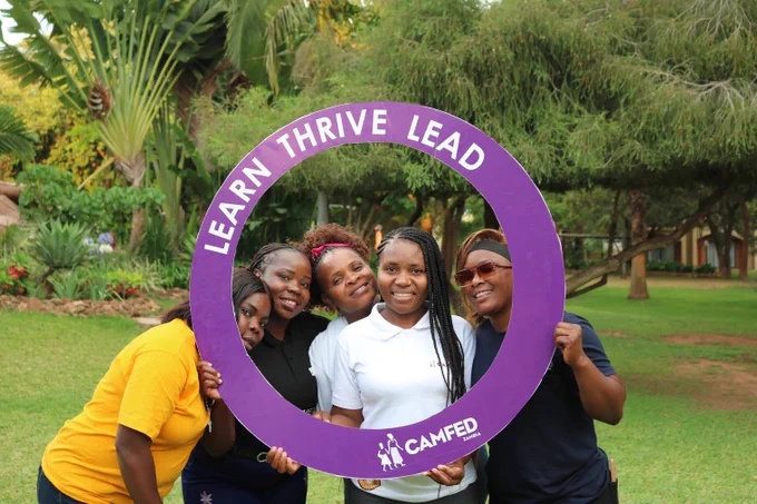 Four individuals posing behind a large purple circular frame with the text "LEARN THRIVE LEAD" from CAMFED, standing in a grassy area with trees in the background.