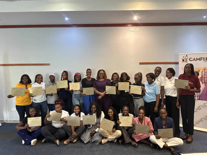Group of individuals holding certificates at a CAMFED event, smiling in front of banners with the organization's logo.