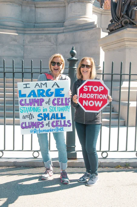 two pro-life activists hold signs that say "stop abortion now" and "I am a large clump of cells standing in solidarity with small clumps of cells"