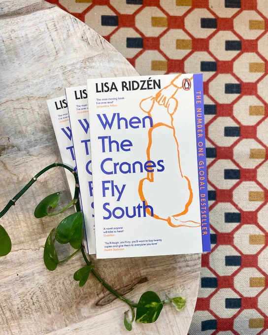 A paperback copy of When the Cranes Fly South by Lisa Ridzén placed on a light wooden table. Two more copies are stacked slightly behind it. A trailing green houseplant lies across the table edge, with a patterned rug in red, blue, and cream visible in the background.