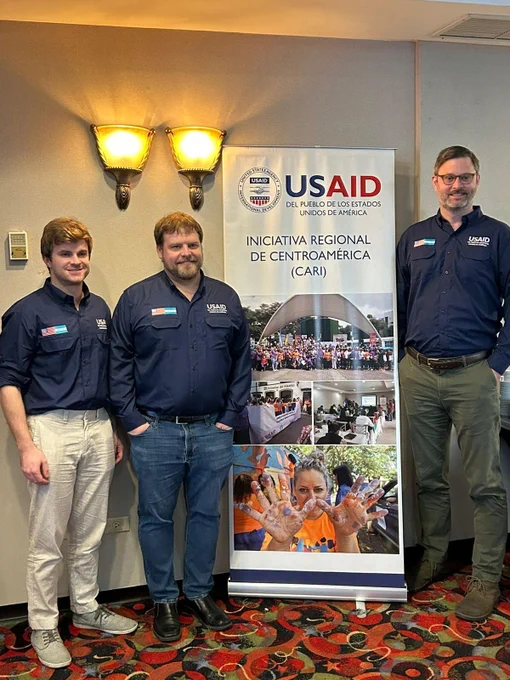 Grady McGinnis, left, with two colleagues in front of a USAID poster that reads "Iniciativa Regional de Centroamerica (CANI)".