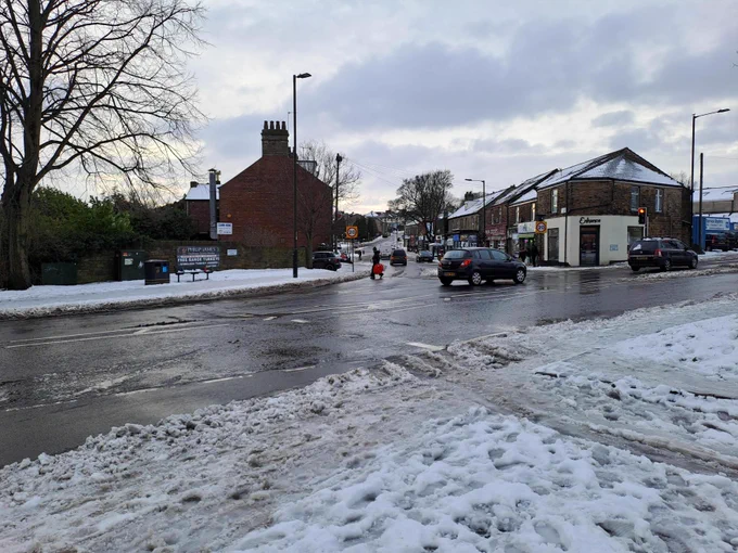 A snowy and slushy scene of Crosspool in Sheffield looking down Sandygate Road towards shops. A person with a sledge is crossing the road and cars drive down the road which is mostly clear of snow.

