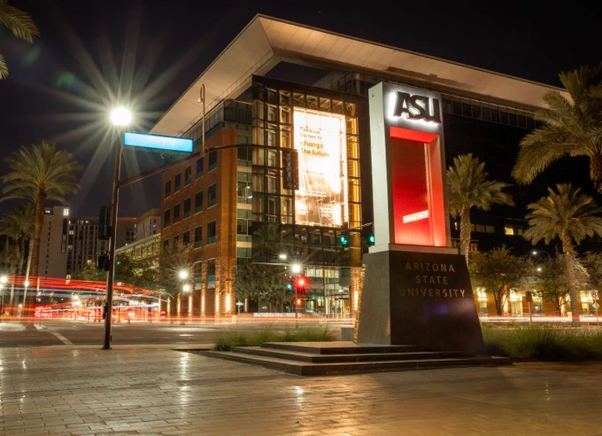 Illuminated Arizona State University sign at night with modern campus building and palm trees in the background.