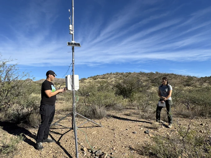 USGS scientists B. Smith and R. Huang stand next to and adjust a system to measure evapotranspiration in an arid land stream channel, surrounded by mesquite shrubs and grassland.