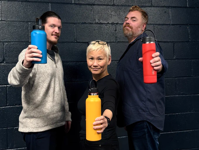 Seth, MoMo, and JW stand in front of a black brick wall and smile as they hold out brightly colored water bottles, Seth holds a blue one, Momo a yellow one, and JW a red one.