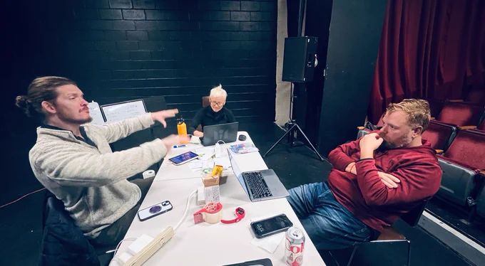 Seth signs MASSAGE as MoMo looks at her laptop and JW looks at Seth in thought. They are seated at a table with papers, tablets, and notes. A music stand in the background with a small whiteboard with notes on it. Black brick wall in the back.