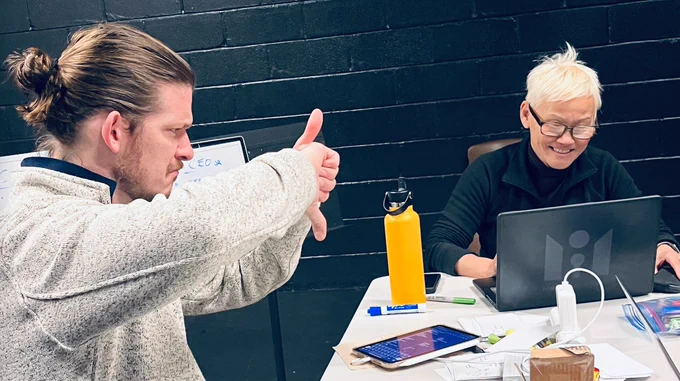 Seth signs “UP AND DOWN” as Momo laughs and looks at her laptop. The table in front of them has papers, tablets, and notes. A music stand in the background with a small whiteboard with notes on it. Black brick wall in the back.
