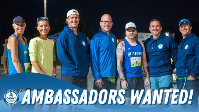 Group of seven Geist Ambassadors in blue shirts standing together under a banner reading 'AMBASSADORS WANTED!' at an outdoor event during nighttime.