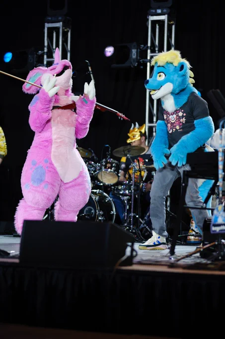 Photo of two fursuiters on a stage - Rhubarb the pink nidoran on the left is looking up with his paws in the air, Key the cyan otter standing next to him.