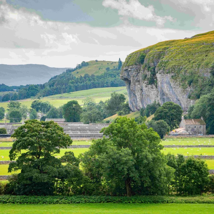 Lush green fields and trees stretch toward a rocky cliff with a small stone house nestled at its base under a cloudy sky.