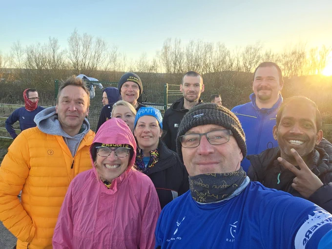 Nine parkrunners, some wearing sportswear and headbands, smiling cheerfully at a parkrun event on a sunny day.
