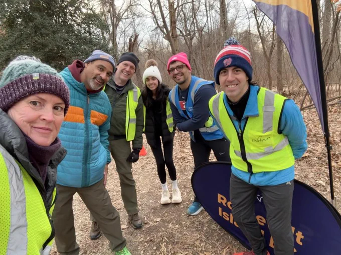 A group of parkrun volunteers posing cheerfully next to the purple parkrun panel at the Roosevelt Island parkrun event. 