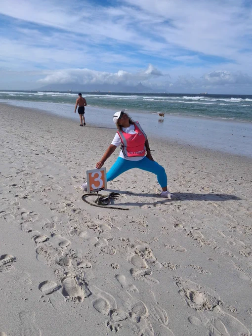 a parkrun volunteer picking up a marker numbered three cheerfully on a sandy beach during a parkrun event, with the ocean and another parkrunner in the background.