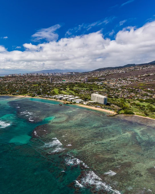Aerial view of Waikiki, showing clear blue waters along the coast and dense urban development extending into verdant hills.