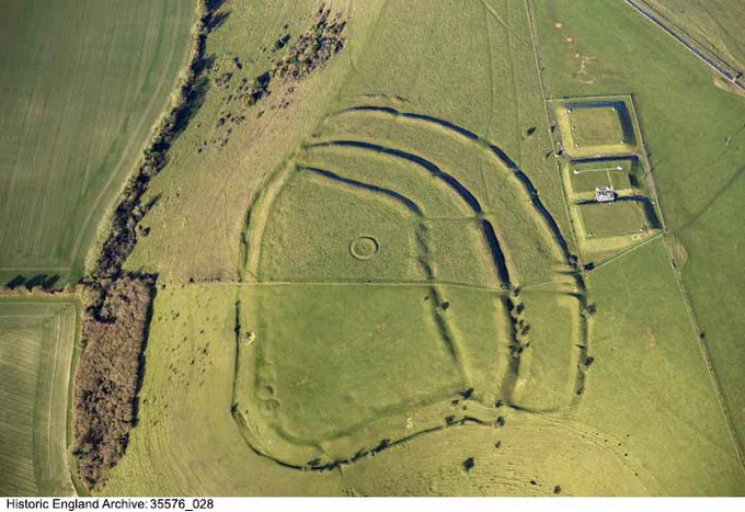 A colour, oblique aerial photo of a large hillfort earthwork with raised banks and ditches. The grass is pale green and yellow. In the centre of the photo is the hillfort earthwork. At its centre is a small circular feature. To the right of the main earthworks are three rectangular areas. A road is just visible running diagonally across the top right corner of the photograph. On the left of the photo is a line of trees and scattered shrubs.