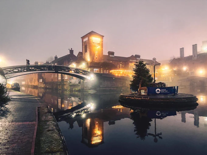 A foggy city scene in Birmingham. Old buildings stand around the canal lit up by street lights. A Christmas tree stands on a canal island and a bridge arches over the still water. 