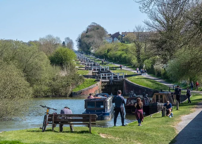 A lock flight stretches upwards into the distance surrounded by trees. Boats float at the bottom of the lock flight while people pass and sit alongside the canal on a bench. 