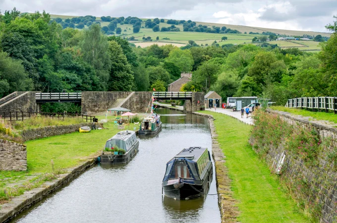 A bright day on a rural canal. Canal boats moor along the towpath and green hills stretch into the distance. Stone and iron bridges stand around the canal.