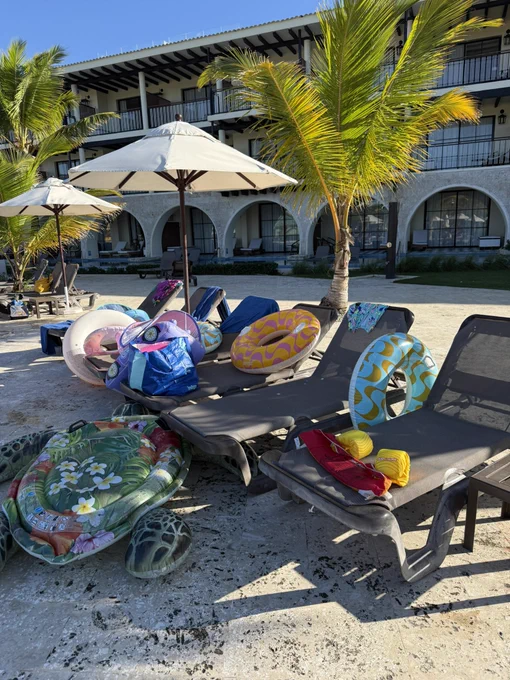 Sunny resort pool deck scene in the Caribbean or Mexico. Multiple black mesh lounge chairs are scattered with bright inflatable pool toys and floats: large green sea turtle, yellow and pink donuts, blue and purple rings, floral turtle float, and several others. Palm trees cast shadows across the stone-tiled area, with a two-story white stucco building featuring arched walkways and black wooden beams visible in the background.