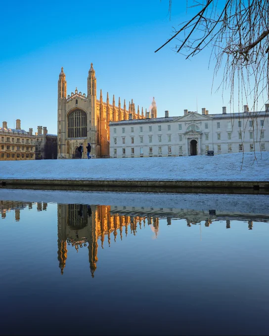 Reflection of King's College Chapel in the River Cam, with a dusting of snow covering the surrounding ground and a clear blue sky.
