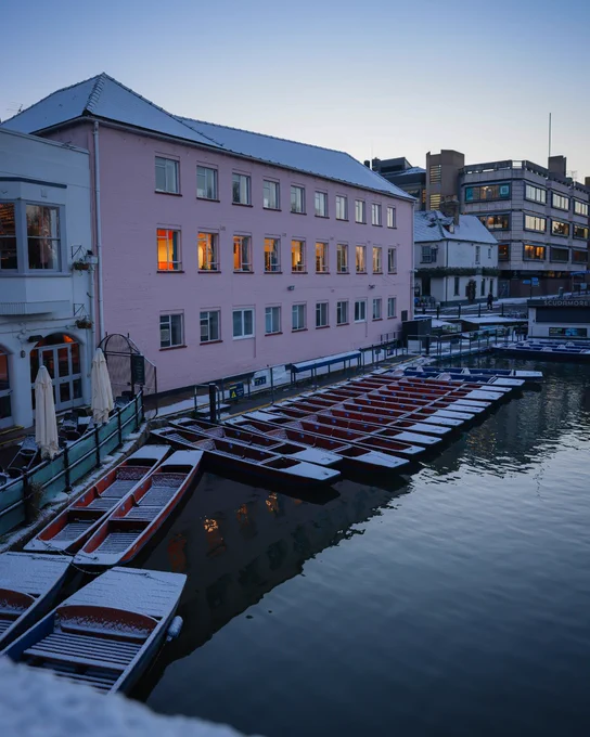 A view of punts lined up on the River Cam in Cambridge on a winter morning, with a pink building illuminated in the background.