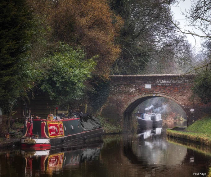 A narrowboat painted in traditional green, red and yellow moors along a rural canal next to an arched red-brick bridge. Further boats can be seen moored along the towpath in the distance, smoke rising from their chimneys.  