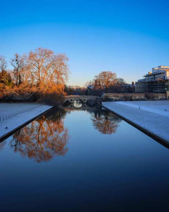 View of the River Cam in Cambridge during winter, showing a frost-covered riverbank and reflected trees with the Clare College Bridge visible in the distance.