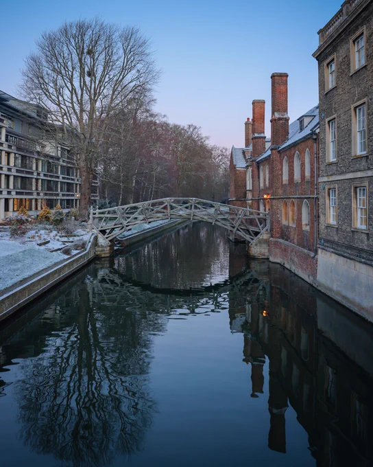 Mathematical Bridge over the River Cam at Queens' College, Cambridge, flanked by historic buildings with snow-dusted roofs on a winter morning.