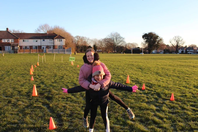 Two junior parkrunners smiling with one carrying the other piggyback, in a grassy field with orange cones, on a sunny day.