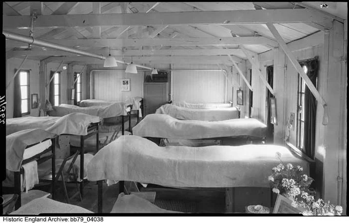 A black and white photo of a dormitory with rows of neatly made beds covered with white sheets. The room has a timber interior. The wooden beams of the roof structure are exposed. In the foreground, on the right, are flowers on a table which is out of view.