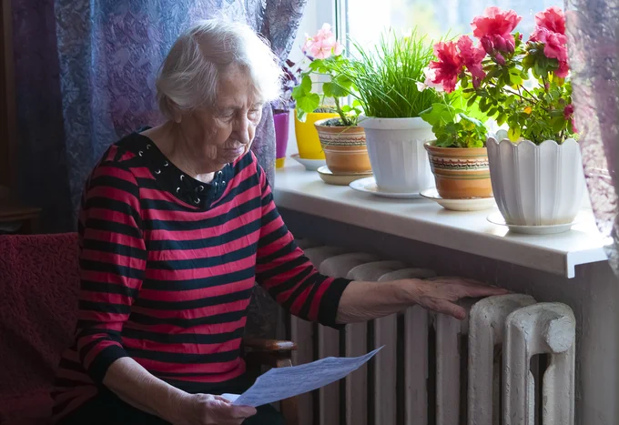 An older woman sat by a radiator reading a bill