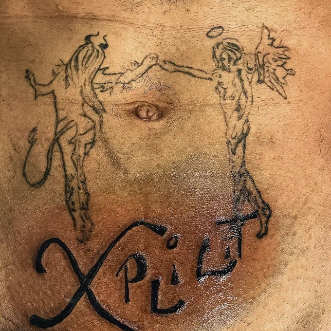 The word Xplicit or “explicit” tattooed on a man’s stomach beneath his belly button. The tattoo also features an angel and demon holding hands with their hands clasping together about the navel of the individual featured in the photograph. The photograph belongs to Thesin Films. 