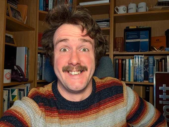 a ridiculous and hairy man wearing a stripy jumper in a book-ridden office space looking baffled but pleased