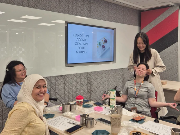 Oracle employees engage in a 'Hands-on Aroma Glycerin Soap Making' activity, as displayed on a screen. One woman massages another's temples, while two others smile at the camera. 