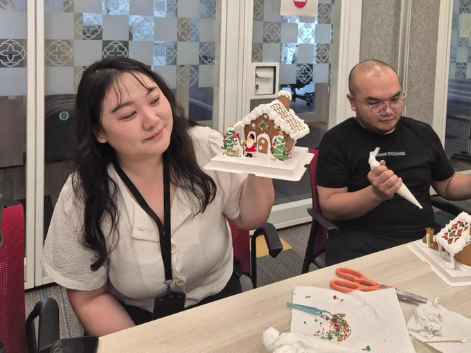 Oracle employee holds up a decorated gingerbread house while a man next to her decorates another one. 
