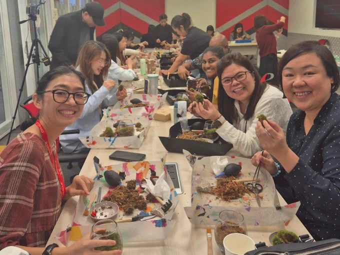 A group of Oracle employees are at a crafting table, smiling. Each person has various craft supplies in front of them, working on assembling terrariums in glass containers.