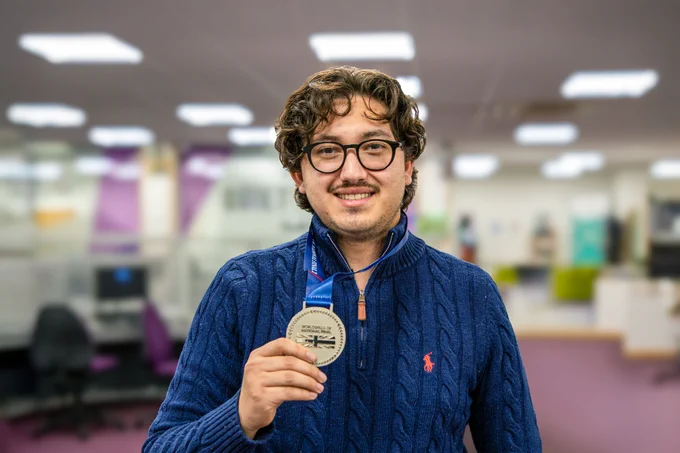 Michael smiling and holding a silver medal. Behind him is a library work space with desks, chairs, a purple carpet and fluorescent lighting. 