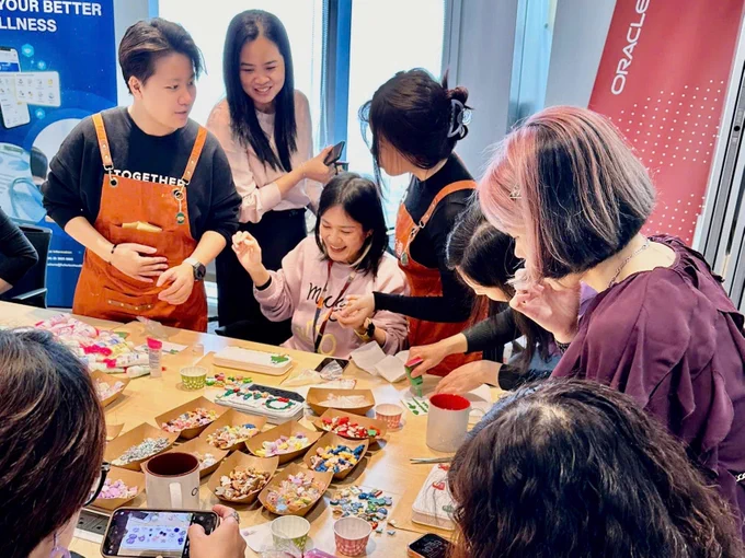 A group of Oracle employees are gathered around a table covered with crafting supplies, including beads, glue, and small containers, creating together in a well-lit room. 