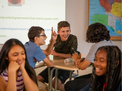 Middle school students actively discussing a classroom topic while seated at desks with a teacher nearby and a map on the wall.