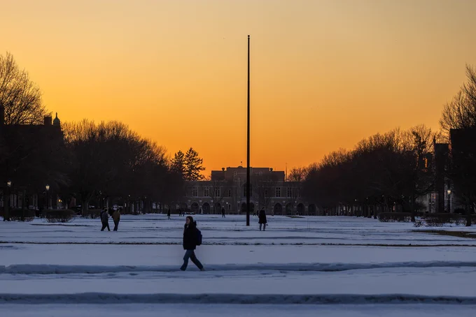 A snowy landscape at dusk, featuring silhouetted figures walking across a snow-covered path. A tall pole stands in the center, with trees lined along the sides and an orange to purple gradient sky in the background.