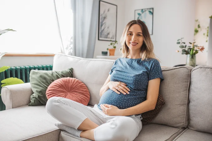 A woman sitting on a sofa in a well-lit living room, holding her pregnant belly. 