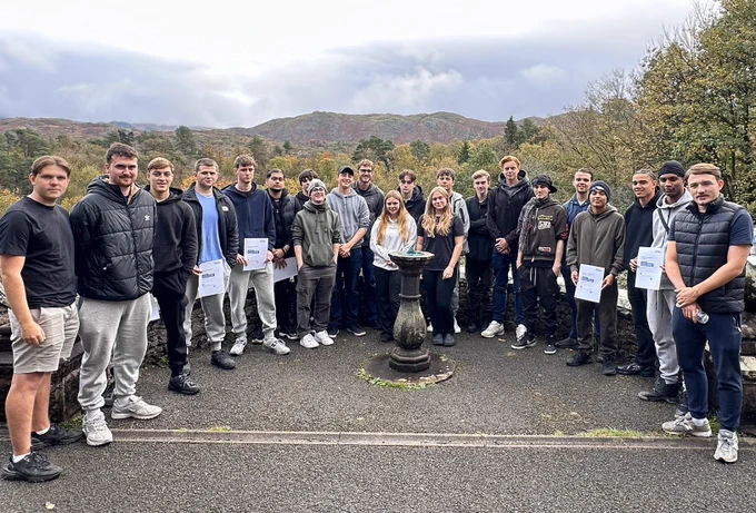 22 apprentices stand at a viewpoint, smiling for the camera. They have been on an Outward Bound Adventure, with a forest landscape behind them. 