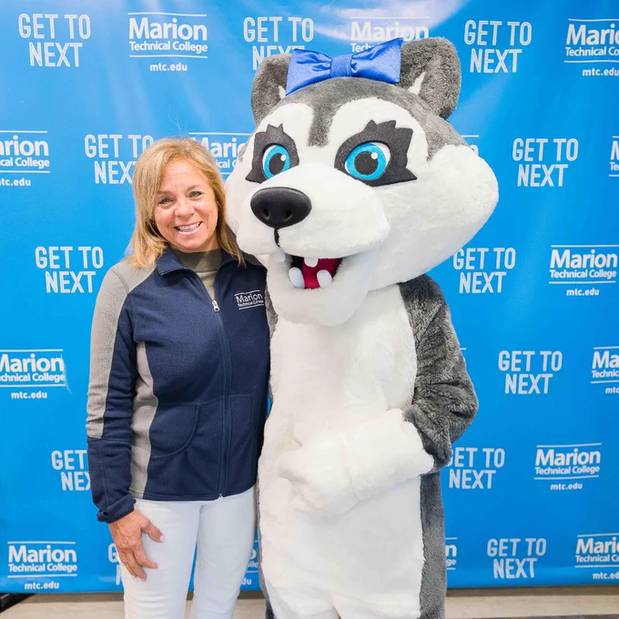 A faculty member poses with one of the college mascots, a giant gray Husky with a white face and blue eyes. 