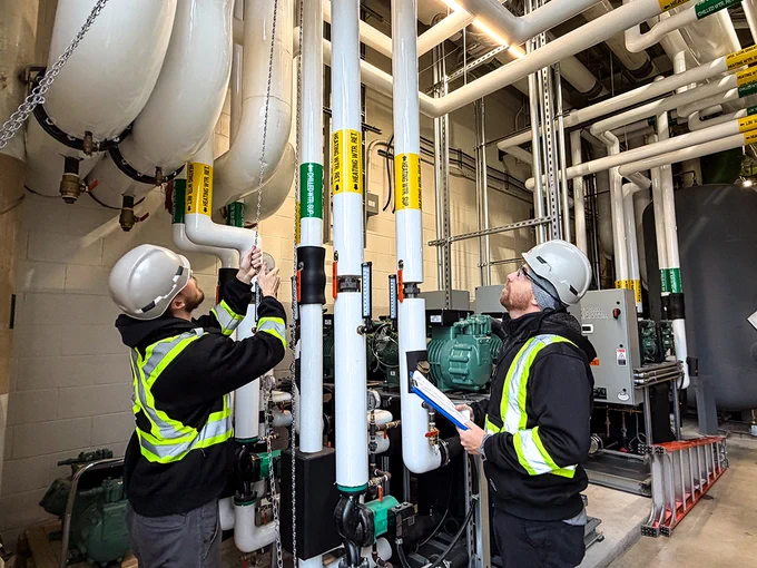 Two people wearing hard hats and high visibility vests are looking at a series of pipes.
