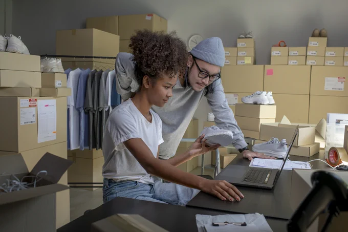 Two people working in a warehouse surrounded by shoe boxes, one holding a sneaker while using a laptop for inventory management.