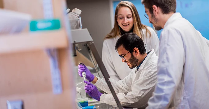 Dr. Anna Blakney is pictured, smiling, while overseeing two of her lab members working under a biosafety cabinet.