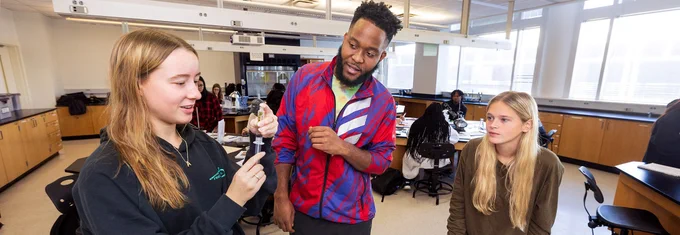 Three East Carolina University students engage in a discussion in a science laboratory, one holding a laboratory instrument while the others observe.
