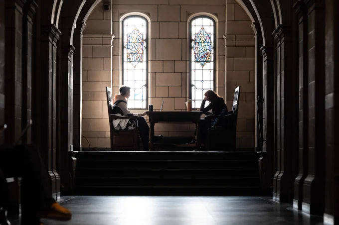 Two individuals are studying at desks in a historic hall with stained glass windows, illuminated by natural light.