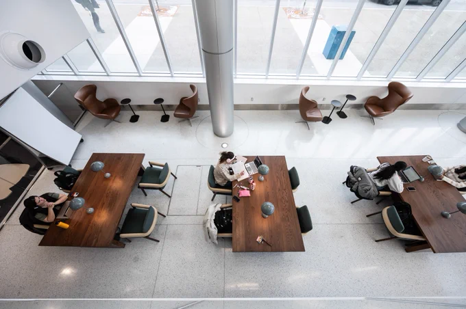 Interior of a modern library with individuals seated at tables, some studying and others using laptops. The setting features contemporary furniture and large windows allowing ample natural light.