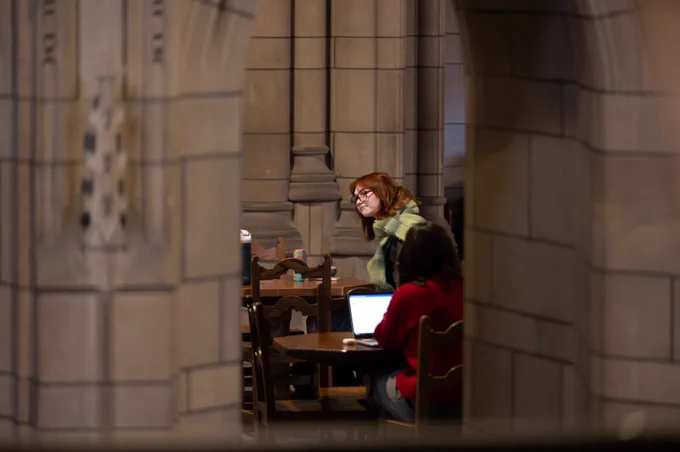 Two individuals are studying at desks inside a historic library, surrounded by stone arches.